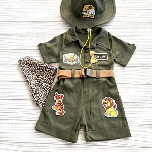 May include: A green safari outfit for a child, featuring a hat, a jumpsuit with a belt, and patches of a tiger, lion, and the words "Safari Tour Guide". The jumpsuit also has a name tag that says "Sevyn".