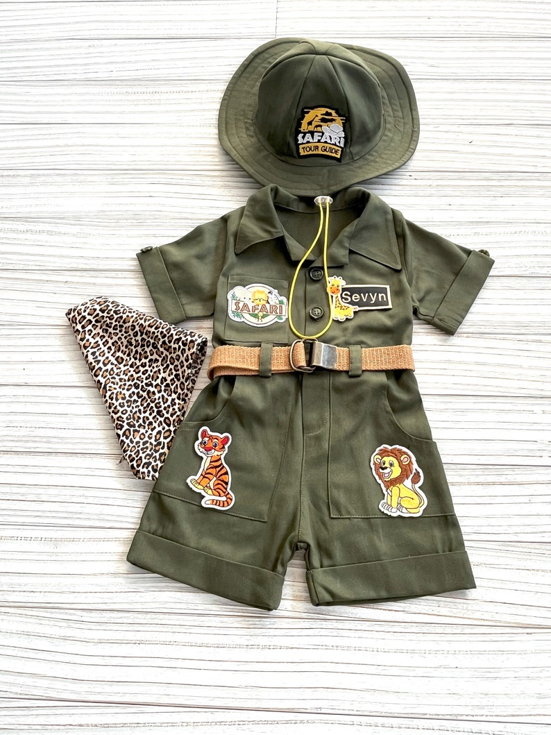 May include: A green safari outfit for a child, featuring a hat, a jumpsuit with a belt, and patches of a tiger, lion, and the words "Safari Tour Guide". The jumpsuit also has a name tag that says "Sevyn".