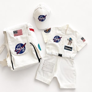 May include: A white NASA-themed outfit set, including a backpack, cap, and shorts set. The backpack and cap feature the NASA logo and American flag patches. The shorts set includes a short-sleeved shirt with the NASA logo and a name tag that says "Sebastian".