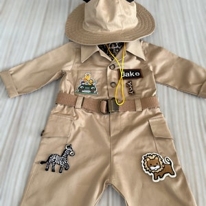 May include: A tan safari costume with a brown belt, a safari hat with black Mickey Mouse ears, and patches of a zebra, lion, and a jeep with giraffes. The costume has a name tag that says "Jake".