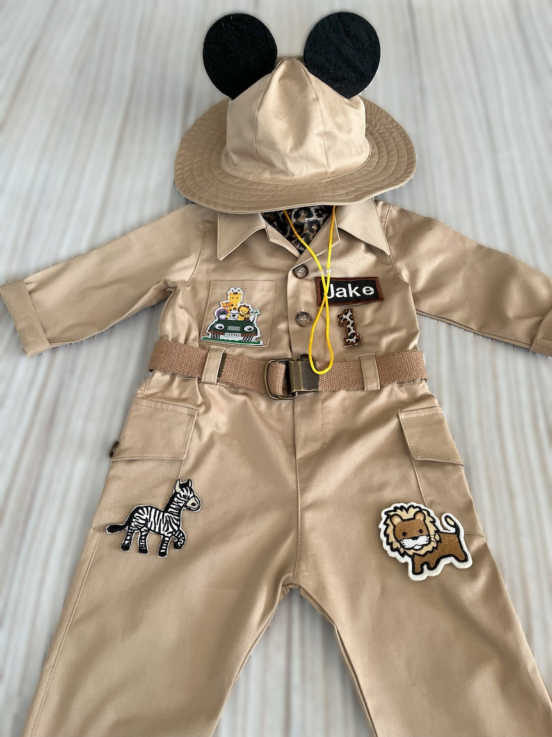 May include: A tan safari costume with a brown belt, a safari hat with black Mickey Mouse ears, and patches of a zebra, lion, and a jeep with giraffes. The costume has a name tag that says "Jake".