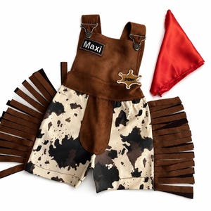 Op de afbeelding: Een bruin en wit cowboy-thema outfit met een rode bandana. De outfit bevat een bruin su&egrave;de-achtig vest met de naam "Maxi" en een sheriff-badge. De shorts hebben een koeienhuidpatroon en bruine franjes.