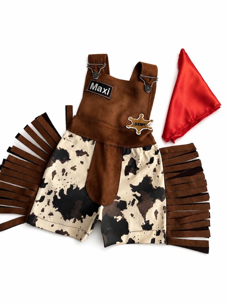 Op de afbeelding: Een bruin en wit cowboy-thema outfit met een rode bandana. De outfit bevat een bruin su&egrave;de-achtig vest met de naam "Maxi" en een sheriff-badge. De shorts hebben een koeienhuidpatroon en bruine franjes.