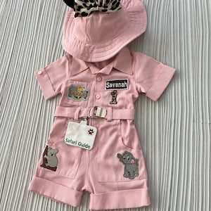 May include: Pink safari outfit for a baby or toddler, featuring a safari hat with black ears and a leopard print bow, a pink safari jumpsuit with pockets and patches, and a safari guide name tag.