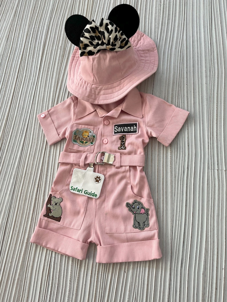 May include: Pink safari outfit for a baby or toddler, featuring a safari hat with black ears and a leopard print bow, a pink safari jumpsuit with pockets and patches, and a safari guide name tag.