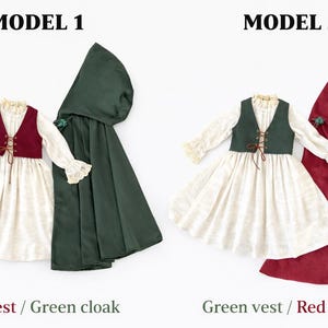May include: Two doll-sized outfits are displayed, each featuring a dress, vest, and cloak. One outfit has a red vest and a green cloak, while the other has a green vest and a red cloak. The dresses are white with long sleeves and lace trim.