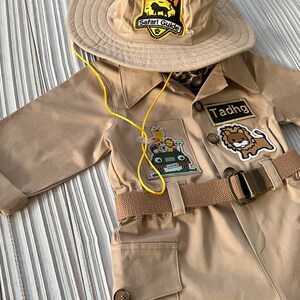 May include: A tan safari jumpsuit with a yellow belt and multiple animal patches. The jumpsuit has a name tag that says "Tadhg". The outfit is topped with a tan safari hat with black Mickey Mouse ears and a patch that says "Safari Guide".