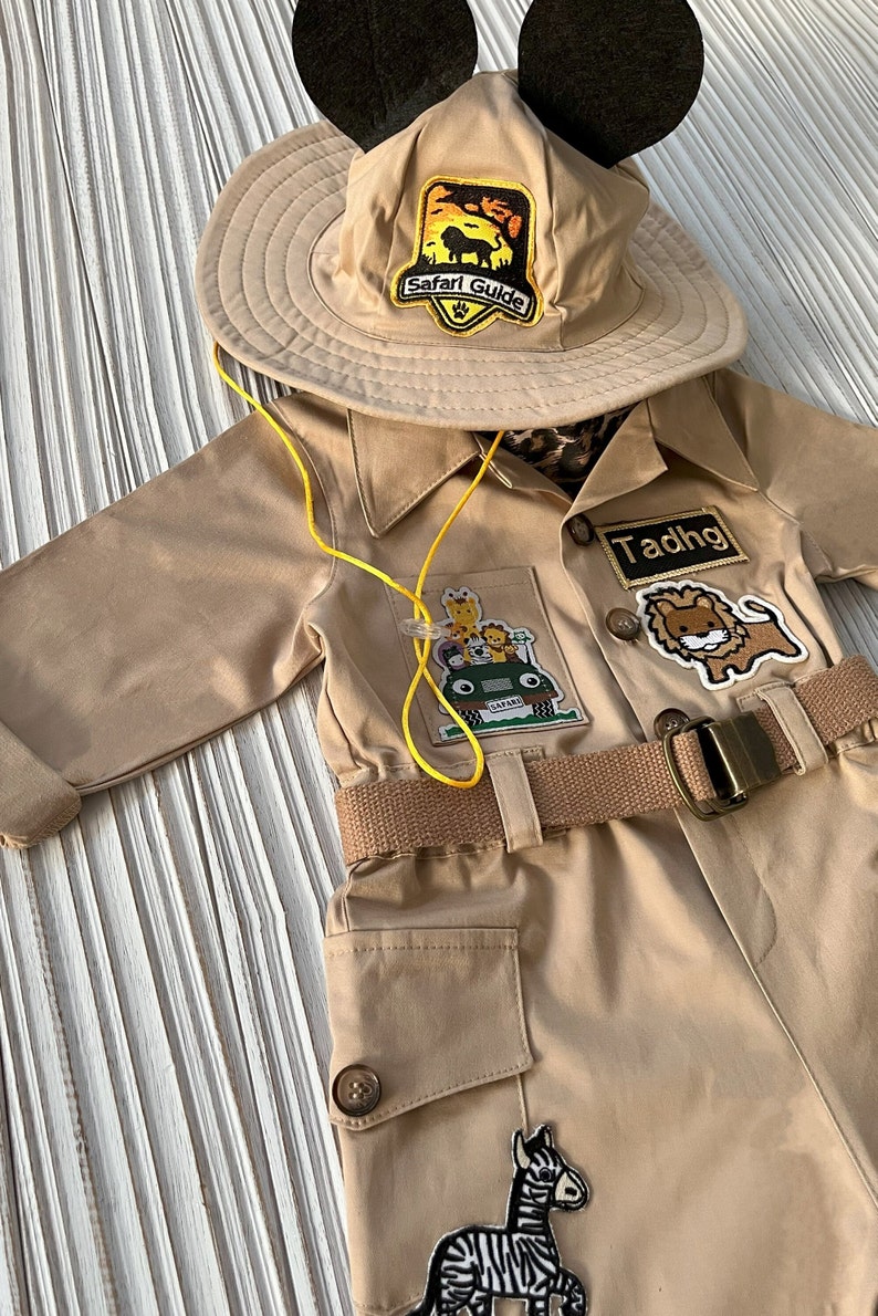 May include: A tan safari jumpsuit with a yellow belt and multiple animal patches. The jumpsuit has a name tag that says "Tadhg". The outfit is topped with a tan safari hat with black Mickey Mouse ears and a patch that says "Safari Guide".