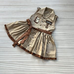 May include: Tan sleeveless dress with a collared neckline, button-down front, and a flared skirt. Features a brown leopard print trim and belt. Includes a patch with a koala and the name "Julian".