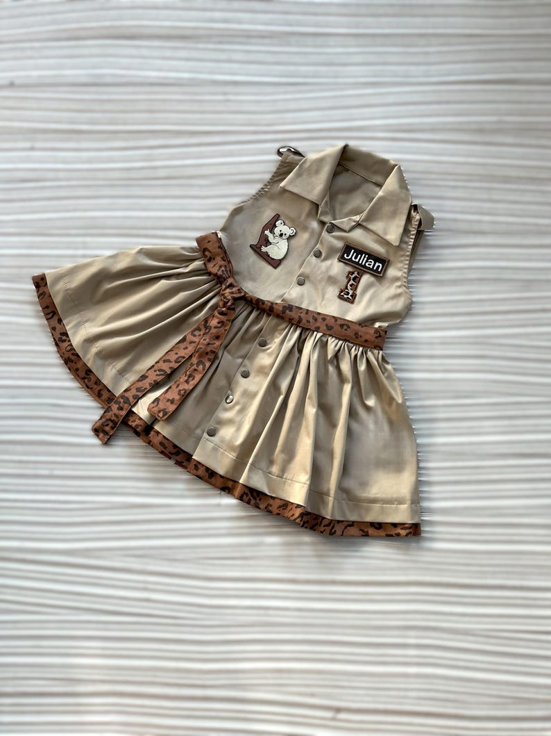May include: Tan sleeveless dress with a collared neckline, button-down front, and a flared skirt. Features a brown leopard print trim and belt. Includes a patch with a koala and the name "Julian".