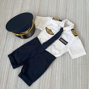 May include: A child's pilot costume set, including a navy blue hat with gold trim, a white short-sleeved shirt with epaulettes and a pilot's wings patch, navy blue shorts, and a black tie. The name "Jaxson" is printed on the shirt.