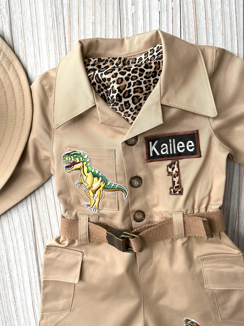 Pu&ograve; includere: Una tuta da safari beige con una toppa di dinosauro, una fodera con stampa leopardata e un'etichetta con il nome "Kailee".