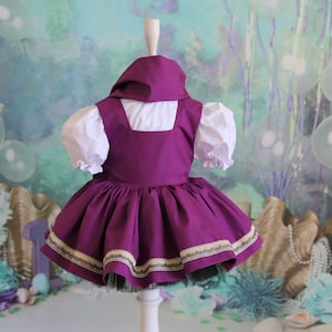 Peut inclure: Une robe violette avec un chemisier blanc et une bordure dor&eacute;e et verte. La robe est sur un mannequin blanc sur un fond vert et bleu.
