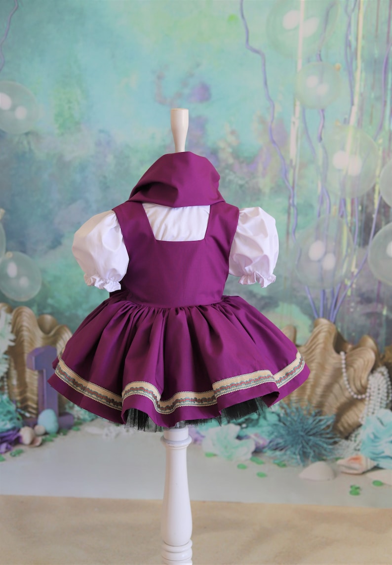 Peut inclure: Une robe violette avec un chemisier blanc et une bordure dor&eacute;e et verte. La robe est sur un mannequin blanc sur un fond vert et bleu.