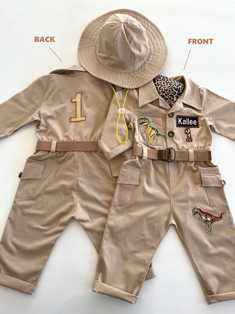 Pu&ograve; includere: Una tuta da safari beige con una toppa di dinosauro sulla gamba destra, un'etichetta con il nome "Kallie" e il numero "1" sul retro. La tuta ha una cintura e due tasche sul davanti. &Egrave; incluso un cappello da safari beige.