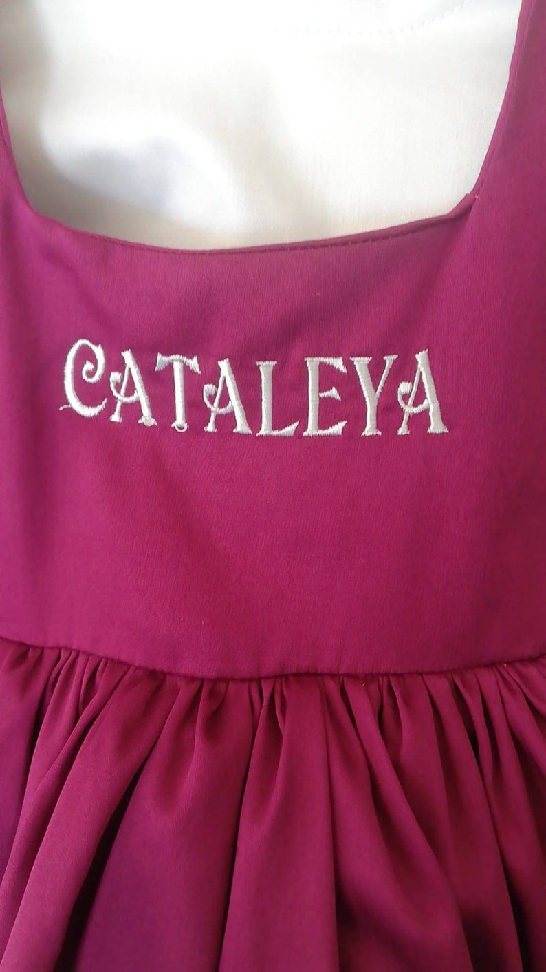 Peut inclure: Une robe bordeaux avec un nom brod&eacute; en blanc "CATALEYA" sur le corsage.