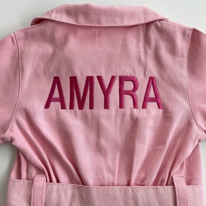 May include: Pink denim jumpsuit with a belt and the name "AMYRA" embroidered on the back in pink.