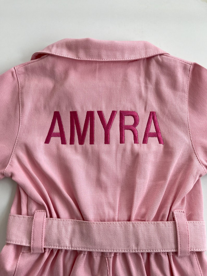 May include: Pink denim jumpsuit with a belt and the name "AMYRA" embroidered on the back in pink.