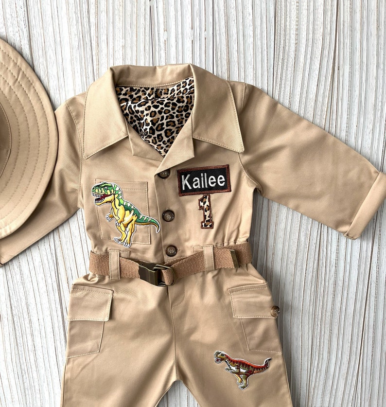 Pu&ograve; includere: Una tuta da safari beige con una cintura marrone, due toppe di dinosauro e un'etichetta con il nome "Kailee".