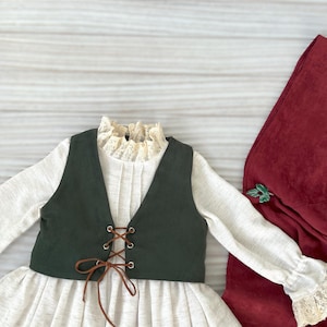 May include: A cream-colored dress with a dark green vest and brown lacing. The dress has long sleeves with lace cuffs and a ruffled lace collar. A burgundy fabric is on the right side of the image.