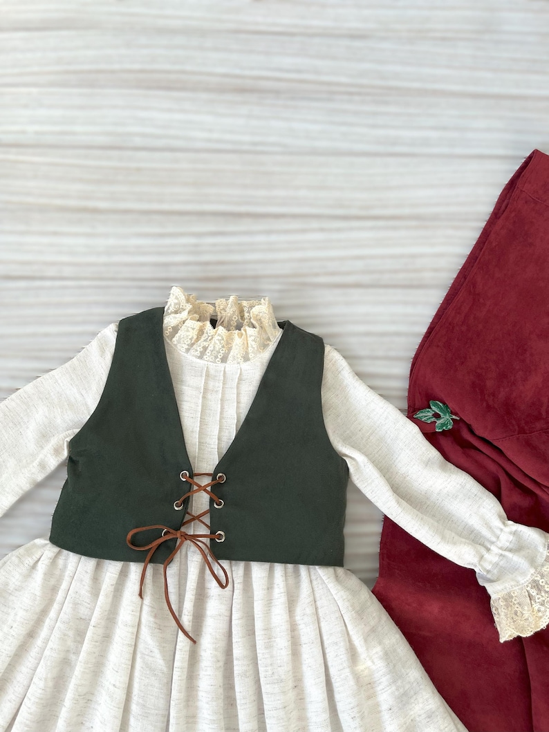 May include: A cream-colored dress with a dark green vest and brown lacing. The dress has long sleeves with lace cuffs and a ruffled lace collar. A burgundy fabric is on the right side of the image.