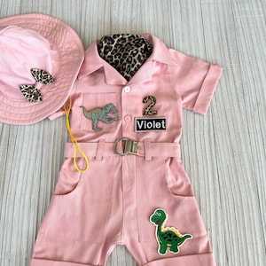 May include: Pink safari-themed jumpsuit with short sleeves, a belt, and two dinosaur patches. The jumpsuit has a name tag that says "Violet" and a number "2" patch. A pink hat with a bow is included.