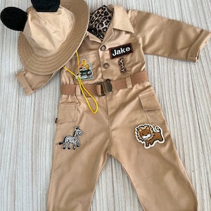 May include: A tan safari jumpsuit with a yellow belt, a black and white zebra patch, a lion patch, and a patch that says "Jake". The jumpsuit has a brown leopard print scarf and a tan hat with black Mickey Mouse ears.