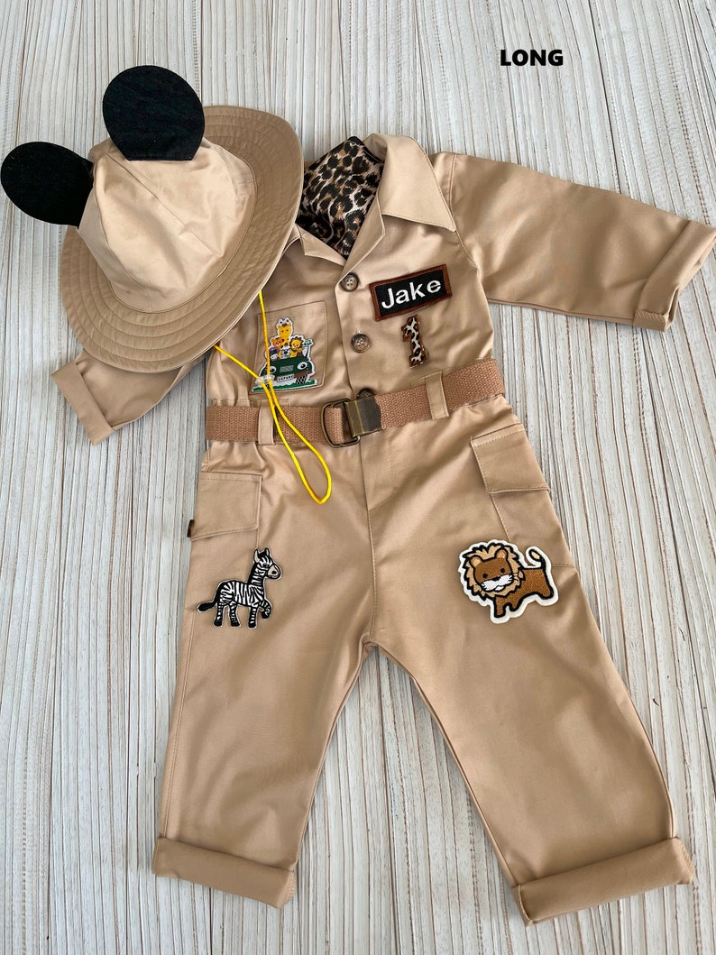 May include: A tan safari jumpsuit with a yellow belt, a black and white zebra patch, a lion patch, and a patch that says "Jake". The jumpsuit has a brown leopard print scarf and a tan hat with black Mickey Mouse ears.