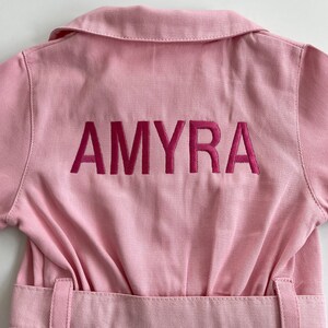 May include: Pink denim jumpsuit with a belt and the name "AMYRA" embroidered on the back in pink.