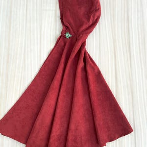 May include: A deep red hooded cloak with a decorative green clasp. The cloak is made of a textured fabric and has a wide, flowing design. The hood is attached to the cloak and is designed to cover the head.