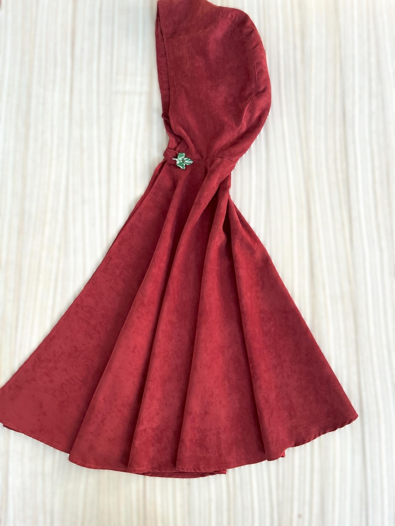 May include: A deep red hooded cloak with a decorative green clasp. The cloak is made of a textured fabric and has a wide, flowing design. The hood is attached to the cloak and is designed to cover the head.