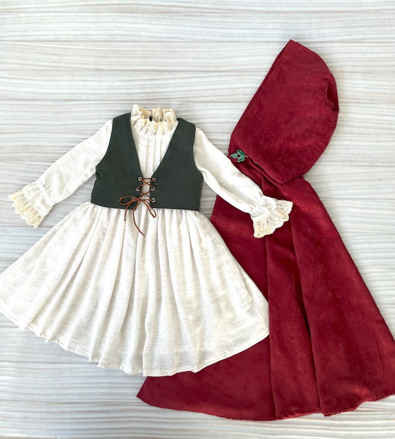 May include: A doll-sized costume set featuring a white dress with a green vest, and a red hooded cape. The dress has long sleeves with lace trim, and the vest has a lace-up front. The cape is a deep red color.