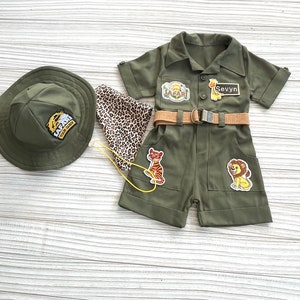 May include: A green safari-themed outfit for a child, featuring a short-sleeved jumpsuit with a brown belt, a safari hat, and a leopard print scarf. The jumpsuit has patches with a tiger, lion, and giraffe, and the text "Safari Tour Guide" and "Sevyvn".