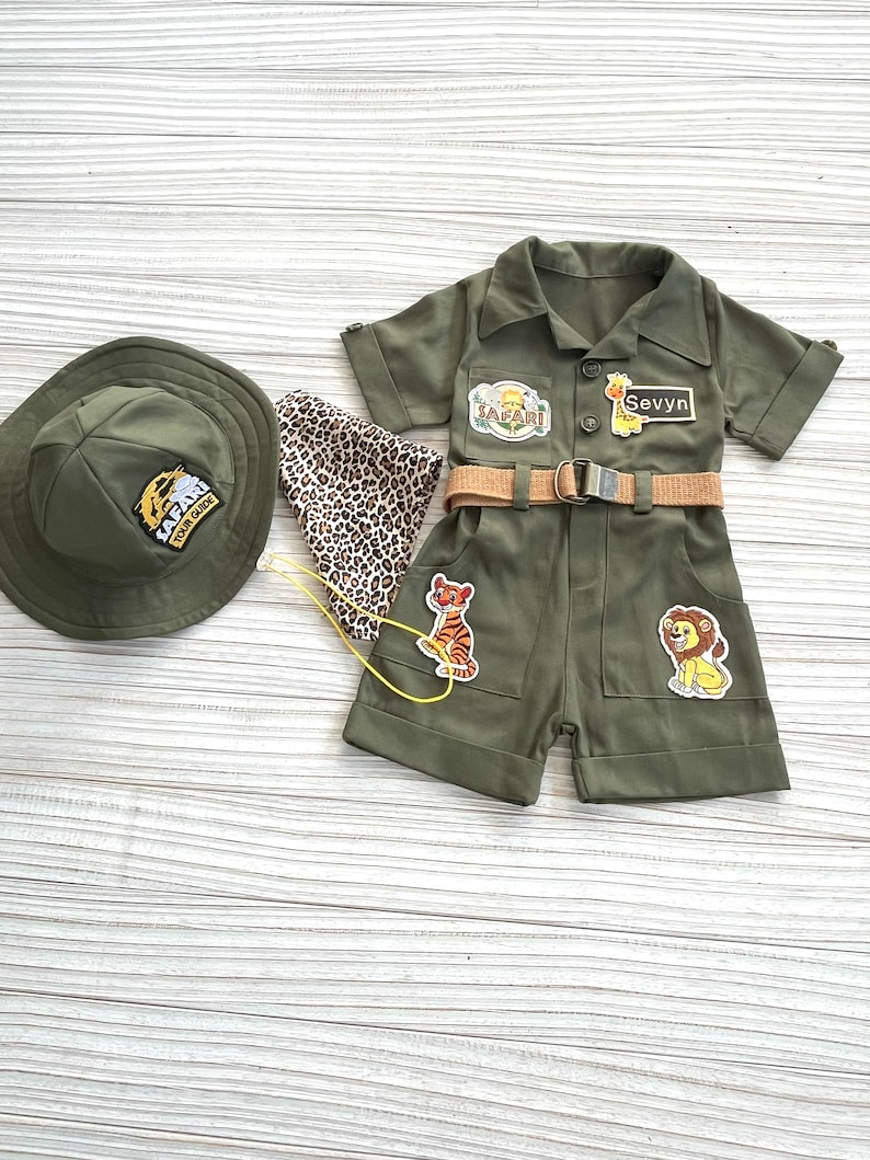 May include: A green safari-themed outfit for a child, featuring a short-sleeved jumpsuit with a brown belt, a safari hat, and a leopard print scarf. The jumpsuit has patches with a tiger, lion, and giraffe, and the text "Safari Tour Guide" and "Sevyvn".