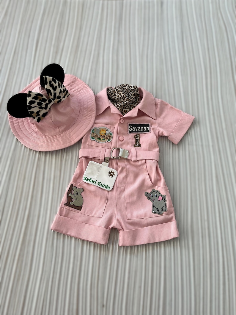 May include: Pink safari-themed outfit for a baby or toddler, featuring a hat with black ears and a leopard print bow, a short-sleeved shirt with a safari patch and a name tag that says "Savannah", and matching shorts with a safari guide patch, a koala patch, and an elephant patch.