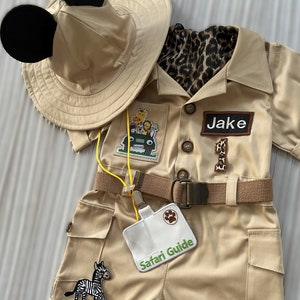 May include: A tan safari outfit for a child with a safari hat with Mickey Mouse ears, a brown belt, and patches of animals and a jeep. The outfit has a name tag that says "Jake" and a badge that says "Safari Guide".