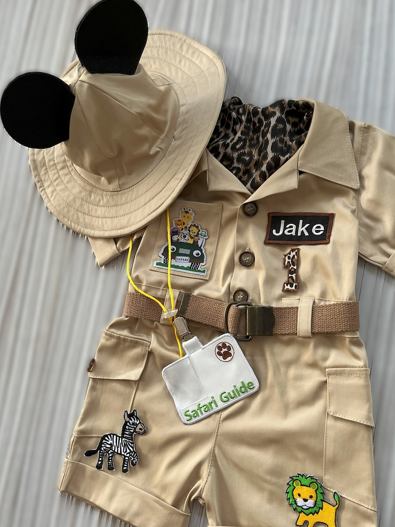 May include: A tan safari outfit for a child with a safari hat with Mickey Mouse ears, a brown belt, and patches of animals and a jeep. The outfit has a name tag that says "Jake" and a badge that says "Safari Guide".