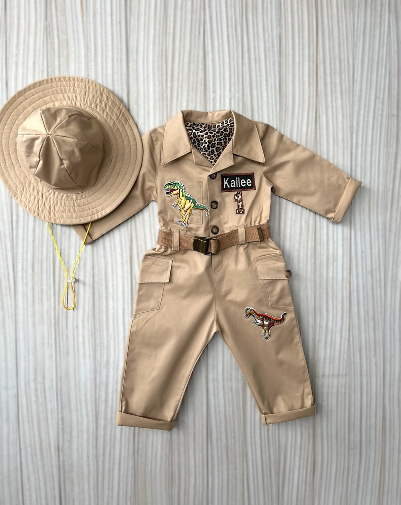 Pu&ograve; includere: Una tuta da safari beige con una toppa di dinosauro, una cintura e un'etichetta con il nome "Kallie". La tuta ha due tasche sul davanti e una toppa di dinosauro sulla gamba destra. La tuta &egrave; abbinata a un cappello da safari beige.