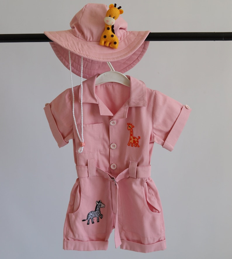 May include: A pink baby jumpsuit with short sleeves and a belt. The jumpsuit has two embroidered giraffes and a zebra. The jumpsuit is paired with a pink sun hat with an embroidered giraffe.