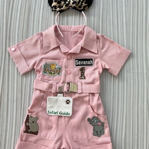 May include: Pink safari jumpsuit with patches, a name tag that says "Savannah", and a badge that says "Safari Guide". The jumpsuit has a belt and pockets. A leopard print headband with black ears is on top.