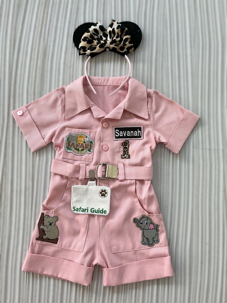 May include: Pink safari jumpsuit with patches, a name tag that says "Savannah", and a badge that says "Safari Guide". The jumpsuit has a belt and pockets. A leopard print headband with black ears is on top.