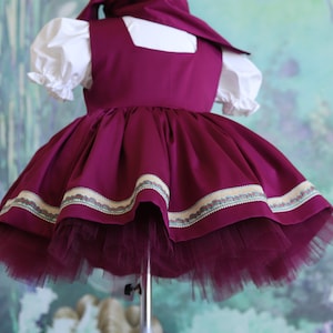 Peut inclure: Une robe bordeaux avec un chemisier blanc et une bordure d&eacute;corative autour de l'ourlet. La robe a une jupe ample et une jupon en tulle.