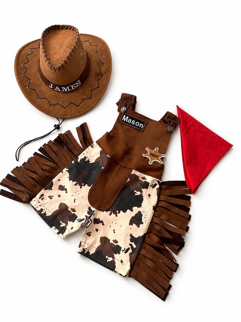 Op de afbeelding: Een bruine cowboyhoed met de naam "JAMES" en een bijpassende outfit. De outfit bevat een bruin vest met een "SHERIFF"-badge, chaps met een koeienhuidpatroon en een rode bandana. De outfit heeft de naam "MASON" erop.
