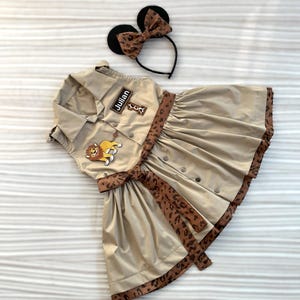 May include: Tan safari-style dress with a pleated skirt, button-up front, and a leopard print belt and trim. Includes a matching headband with black mouse ears and a leopard print bow. The dress has a patch that says "Julian".