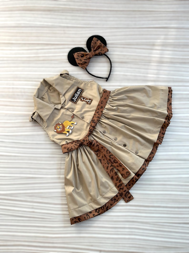 May include: Tan safari-style dress with a pleated skirt, button-up front, and a leopard print belt and trim. Includes a matching headband with black mouse ears and a leopard print bow. The dress has a patch that says "Julian".