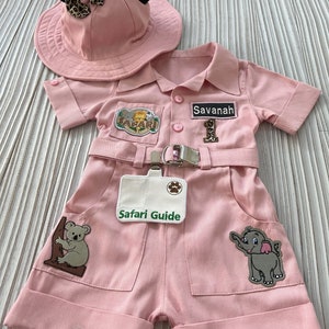 May include: Pink safari outfit for a baby or toddler, featuring a safari hat with black Mickey Mouse ears, a pink jumpsuit with patches, and a name tag that says "Savannah".