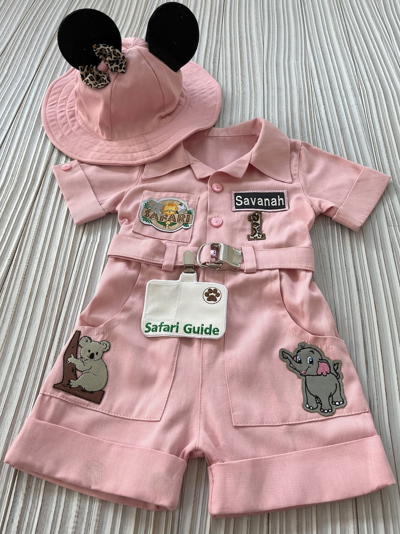 May include: Pink safari outfit for a baby or toddler, featuring a safari hat with black Mickey Mouse ears, a pink jumpsuit with patches, and a name tag that says "Savannah".