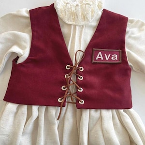 May include: A burgundy vest with a lace-up front and the name "Ava" embroidered on a patch. The vest is layered over a cream-colored blouse with a ruffled collar, creating a vintage or historical aesthetic. The outfit appears to be for a child.