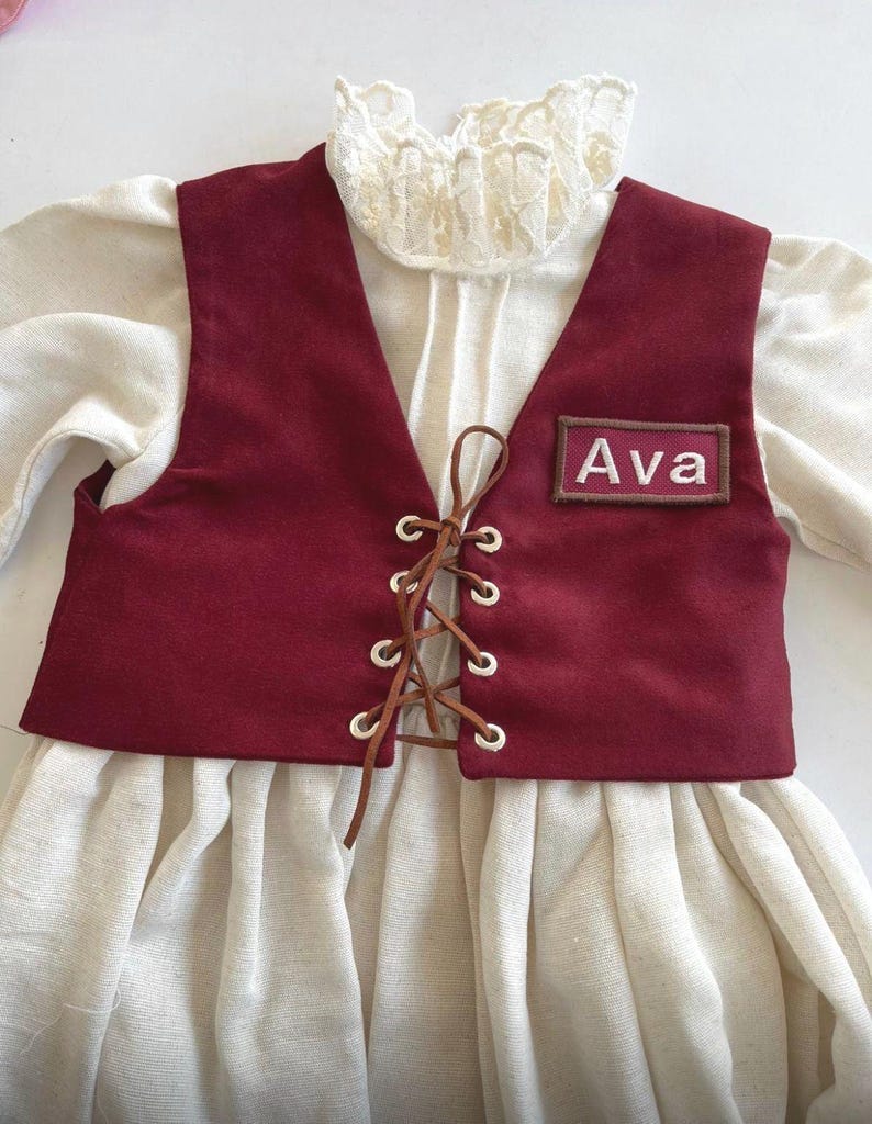 May include: A burgundy vest with a lace-up front and the name "Ava" embroidered on a patch. The vest is layered over a cream-colored blouse with a ruffled collar, creating a vintage or historical aesthetic. The outfit appears to be for a child.