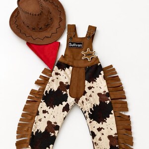 Op de afbeelding: Een bruine cowboyhoed, een rode bandana en een jumpsuit met koeienprint, franjes en een sheriff-badge. Op de jumpsuit staat de naam "Sullivan". Deze western-outfit is perfect voor een kostuum.